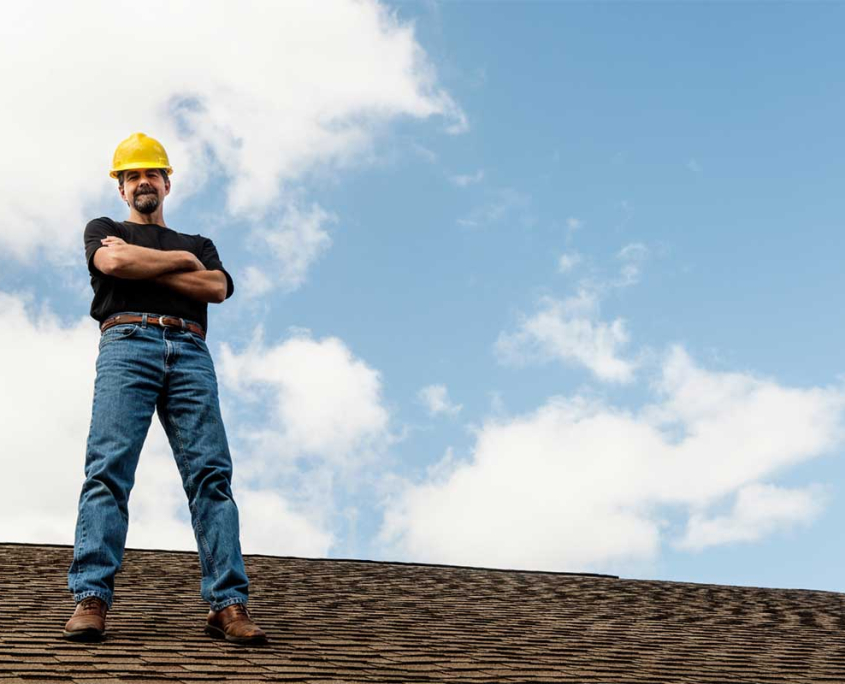 A man stands confidently on the roof of a house, overlooking the surroundings with a thoughtful expression.